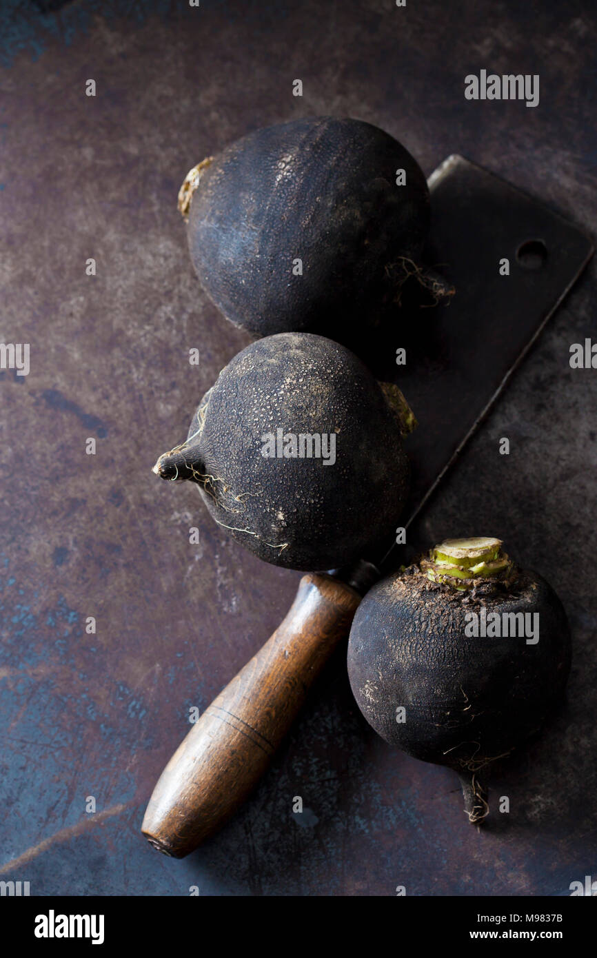 Three black radishes and an old cleaver on rusty ground Stock Photo