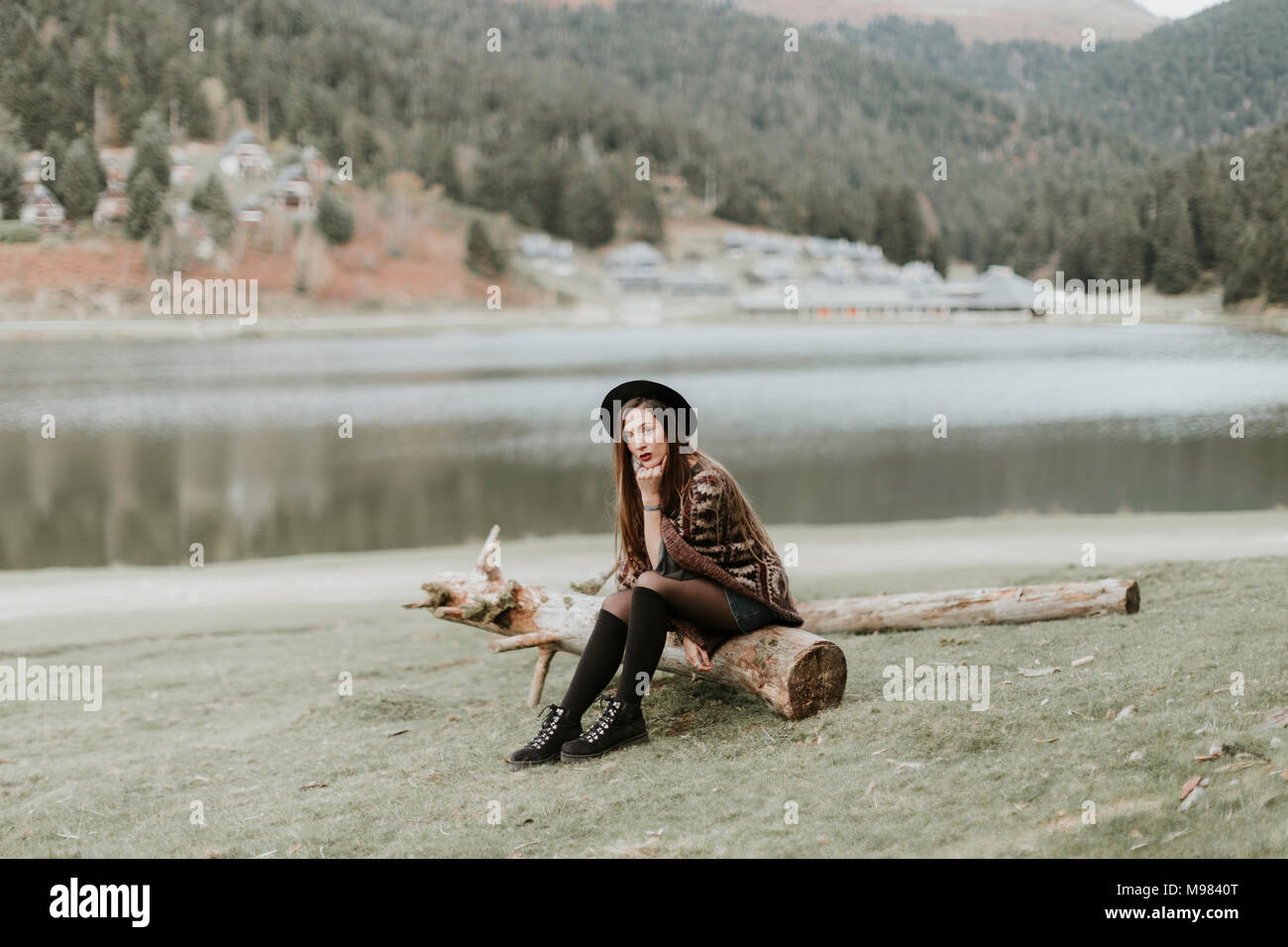 Fashionable young woman wearing hat and poncho sitting on log on a meadow Stock Photo