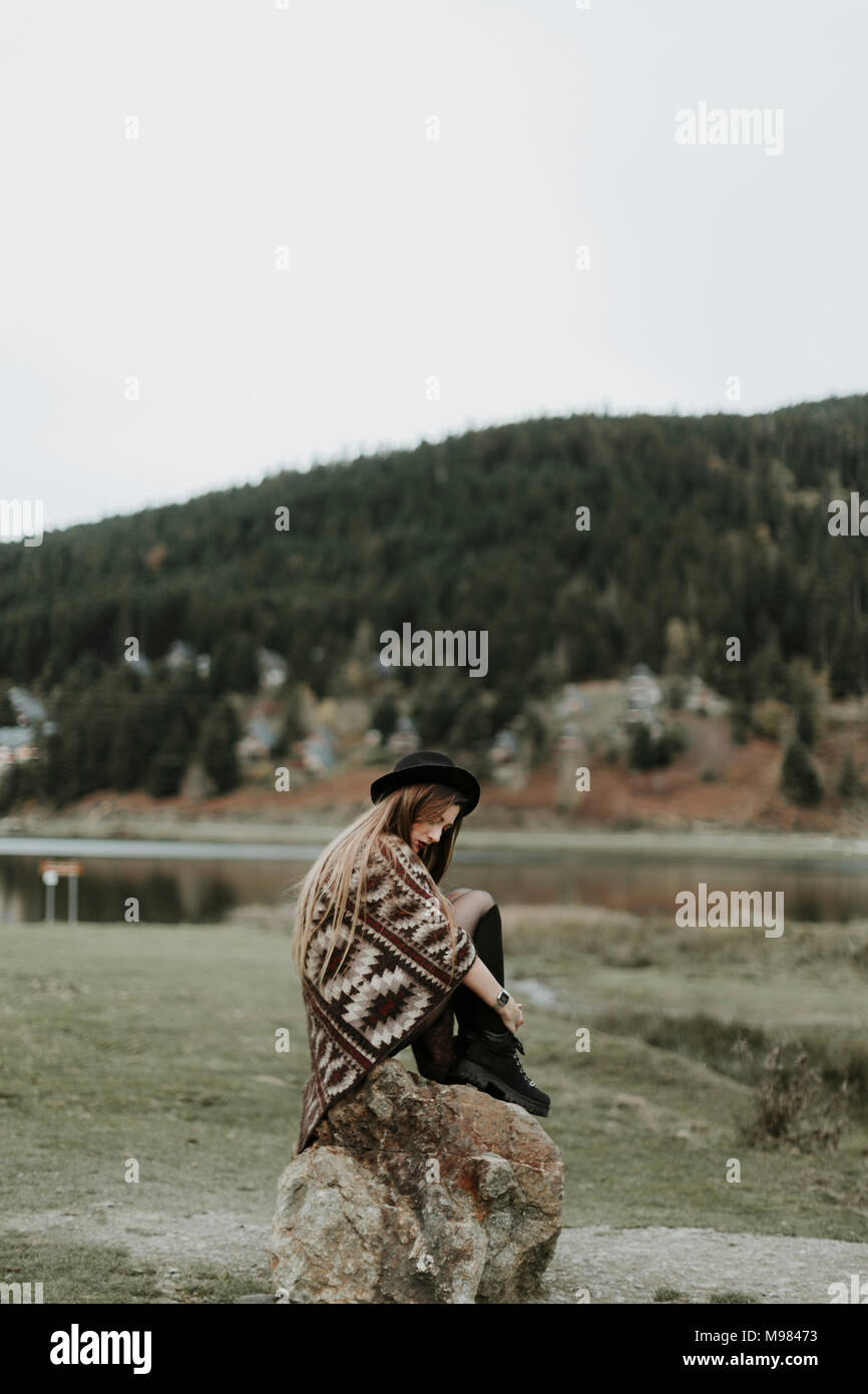 Fashionable young woman wearing hat and poncho sitting on a rock Stock Photo