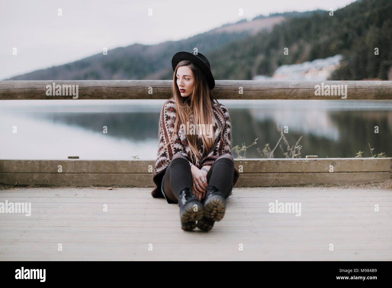 Portrait of fashionable young woman wearing hat and poncho sitting on wood bridge Stock Photo