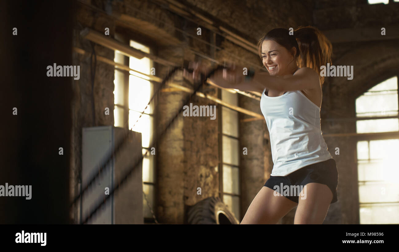 Athletic Female in a Gym Exercises with Battle Ropes During Her Cross Fitness Workout/ High-Intensity Interval Training. She's Muscular and Sweaty Stock Photo