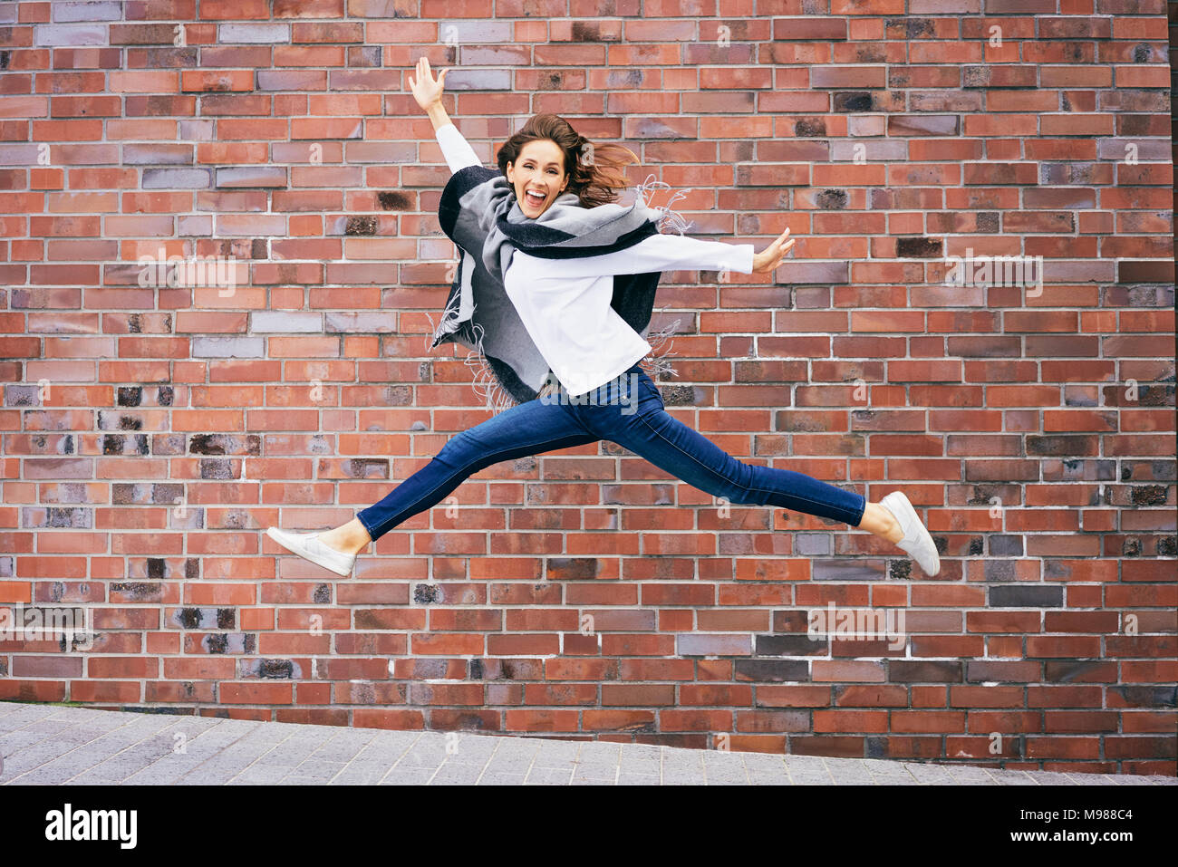 Happy young woman jumping in the air in front of brick wall Stock Photo