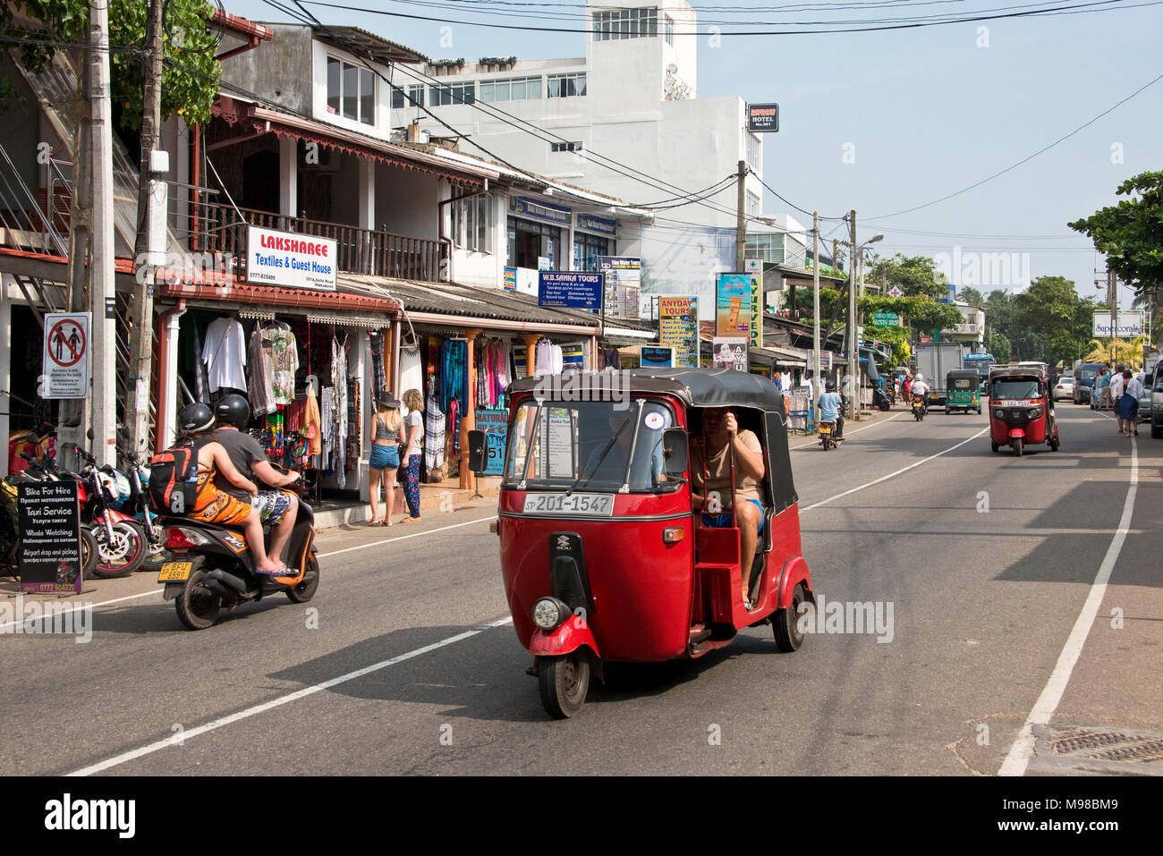 The main tourist area street road in Hikkaduwa, Sri Lanka on a sunny day with blue sky, tuk tuks and scooters. Stock Photo
