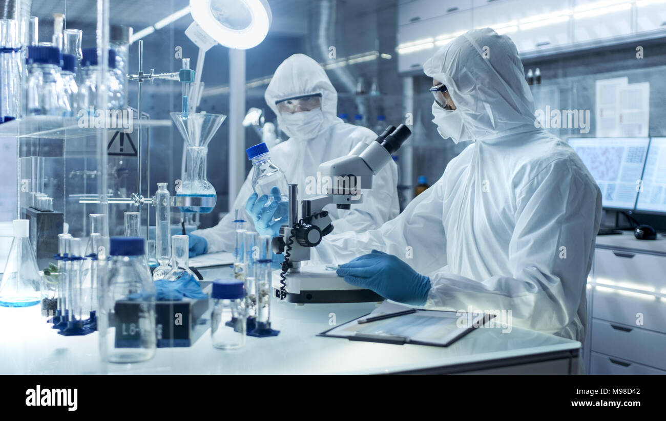 In a Secure High Level Laboratory Scientists in a Coverall Conducting a Research. Chemist Adjusts Samples and Discuss Experiment with Collegue. Stock Photo