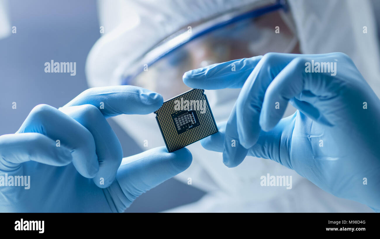 In Ultra Modern Electronic Manufacturing Factory Design Engineer in Sterile Coverall Holds Microchip with Gloves and Examines it. Stock Photo