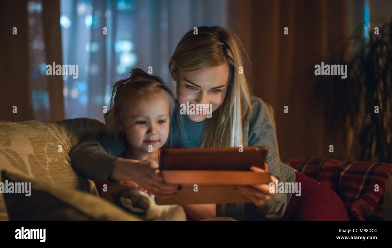 Beautiful Mother and Her Little Daughter are Sitting on a Sofa in the Living Room, They Use Tablet Computer. It's Evening, Room is Cozy and Warm. Stock Photo