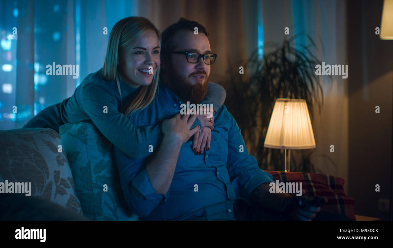 In the Evening Man Sitting on a Sofa Playing Videogames, His Spouse Comes in and Hugs Him. Stock Photo