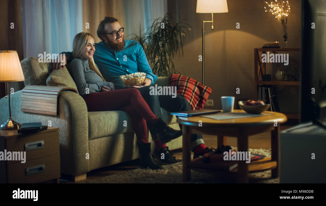 In the Evening Man and Woman are Sitting on the Sofa Watching TV and Eating Popcorn. Living Room is Cozy. Stock Photo