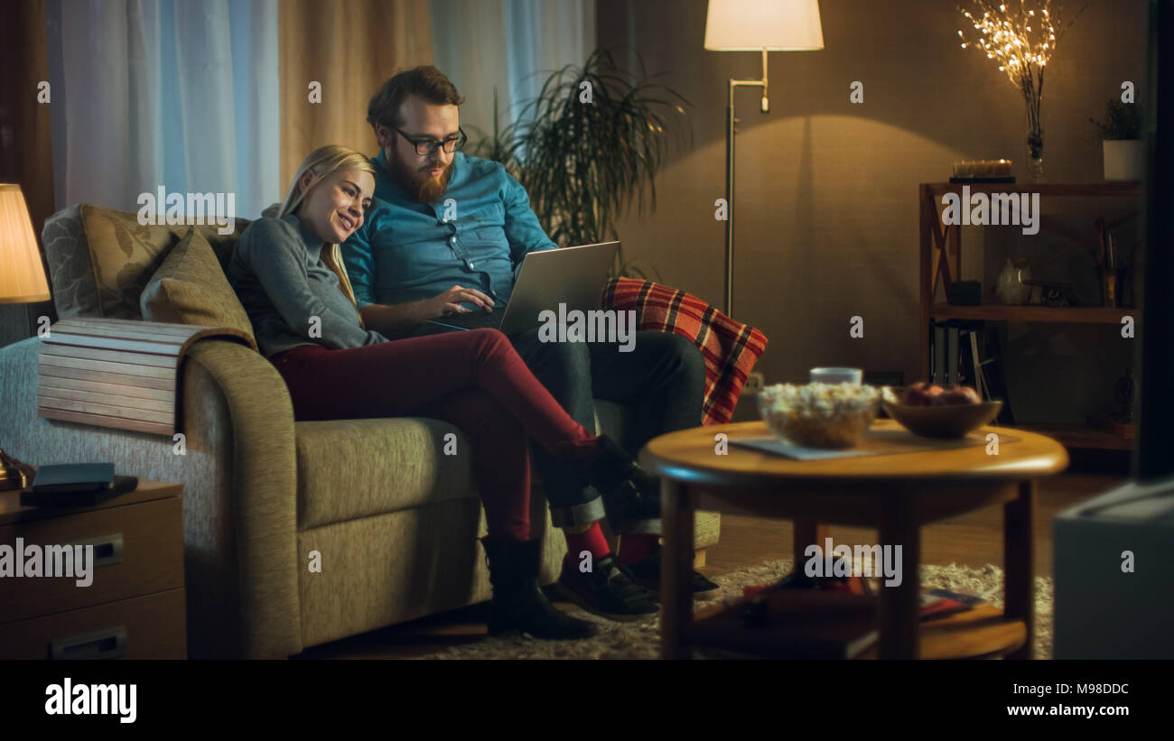 Girlfriend and Boyfriend Sitting on a Sofa with Laptop on His Knees, Watching Videos and Having Fun. Living Room is Cozy. It's Evening. Stock Photo