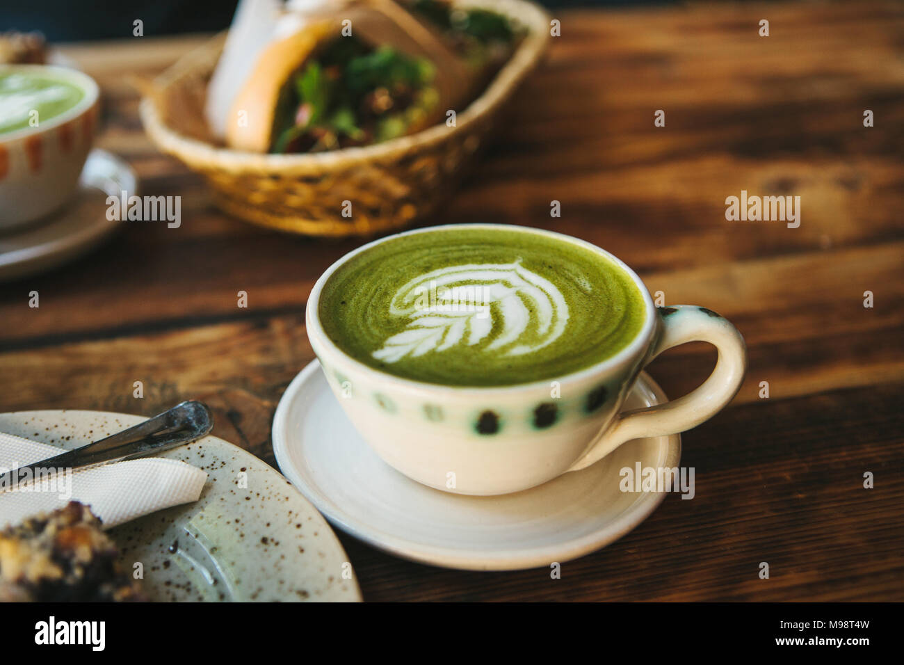 Close-up - ceramic cup with green tea with milky foam with beautiful pattern on the background of blurry basket with food Stock Photo