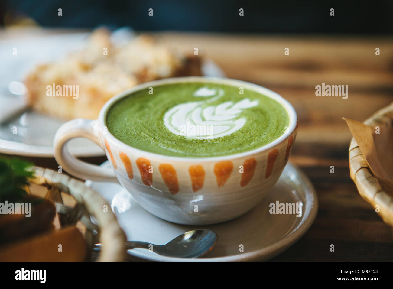 Close-up - ceramic cup with green tea with milky foam with beautiful pattern on the background of blurry basket with food Stock Photo