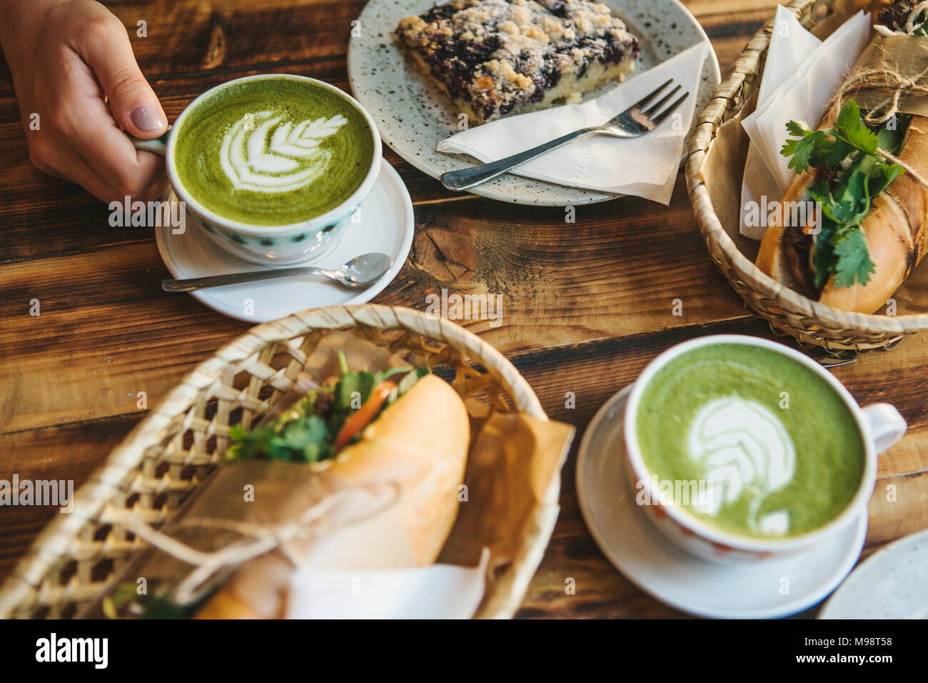 Top view. The girl sitting in cafe and holding mug with green tea with milk next to piece of sweet pie and two sandwiches with vegetables Stock Photo