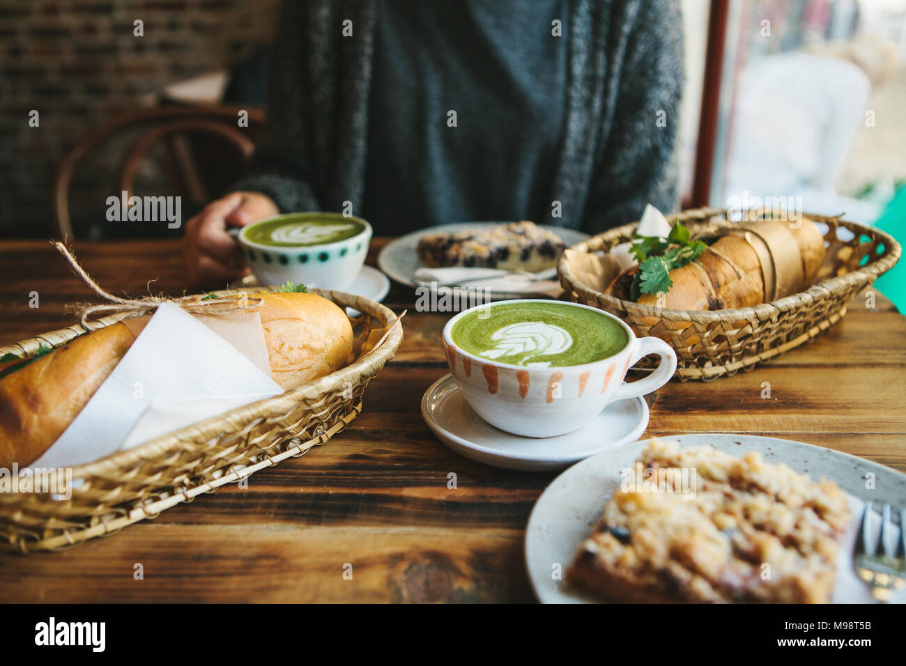 The girl sitting in cafe and holding mug with green tea with milk next to piece of sweet pie and two sandwiches with vegetables Stock Photo