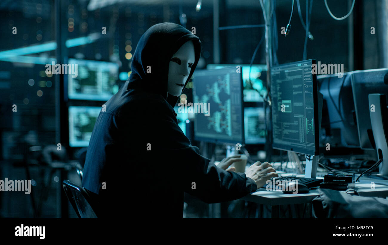 Masked Hacktivist Organizes Massive Data Breach Attack on Corporate Servers. He is in Underground Secret Location Surrounded by Displays and Cables. Stock Photo