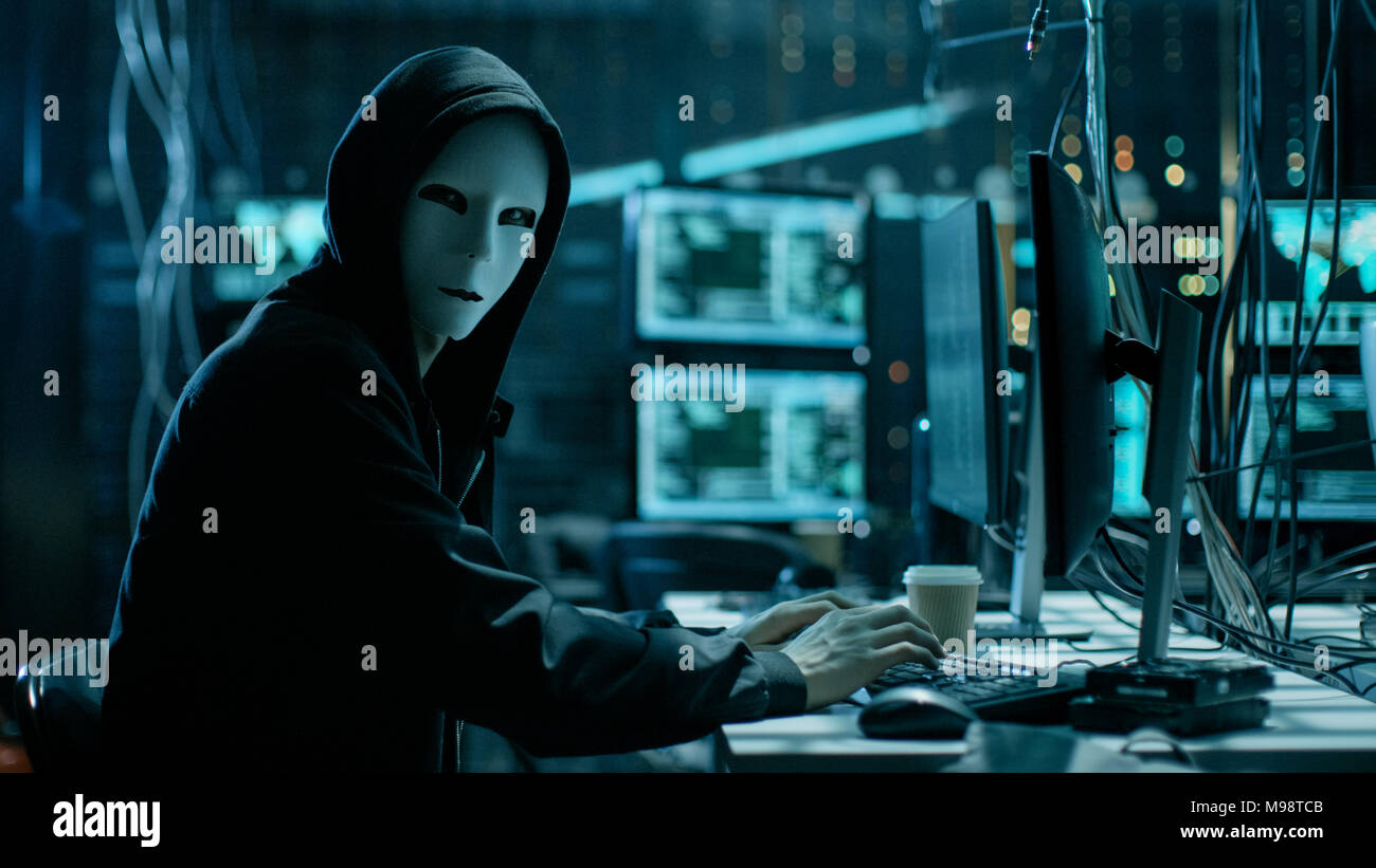 Masked Hacker is Using Computer for Organizing Massive Data Breach Attack on Corporate Servers. They're in Underground Secret Location Stock Photo