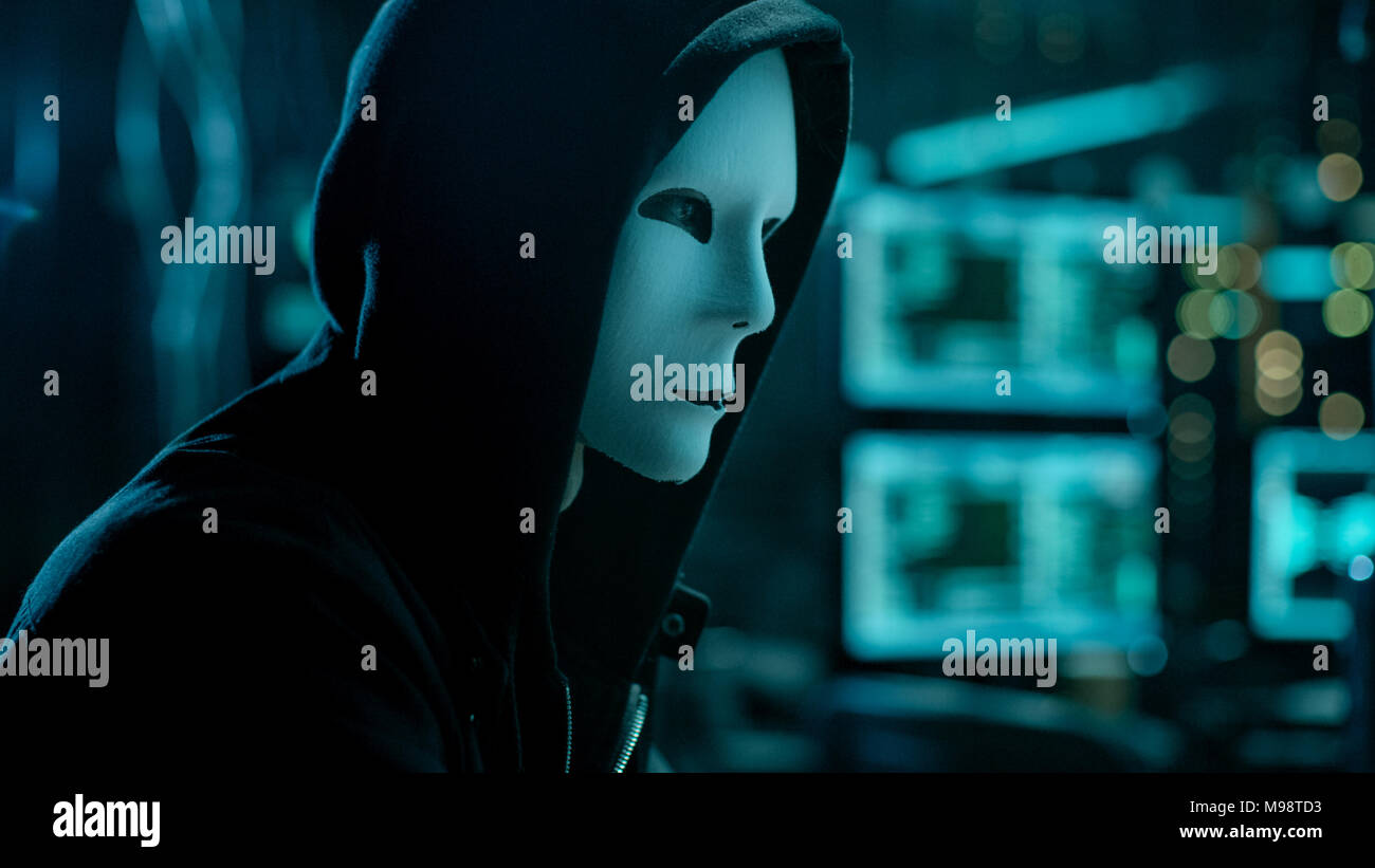Masked Hacktivist Organizes malware Virus Attack on Global Scale. Hacker in Underground Secret Location Surrounded by Displays and Cables. Stock Photo