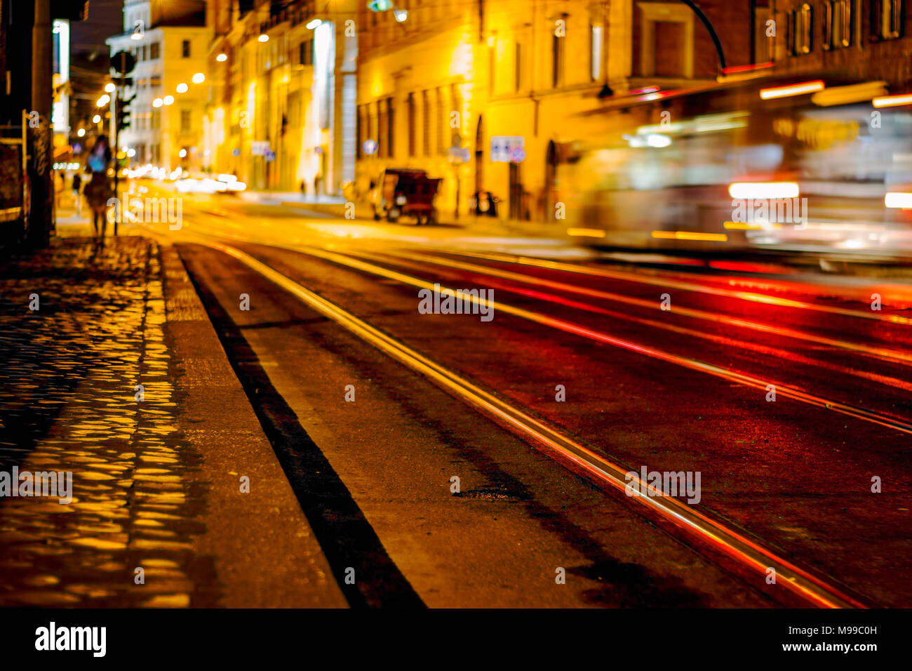 Night scene of Rome featuring a speeding bus with light trails and lights reflecting on tram lines and with motion blur and lone female figure Stock Photo
