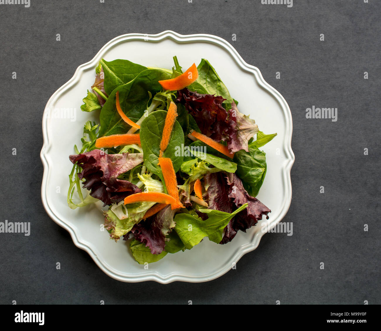 Unseasoned organic mixed greens salad  with carrots in white plate bowl, on dark background, top view-diet food concept Stock Photo