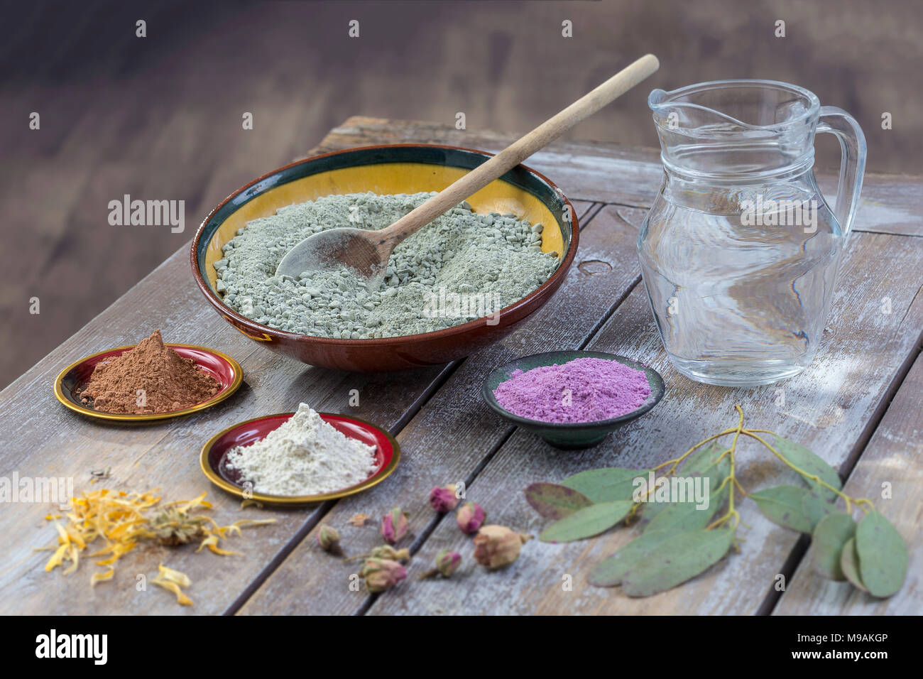 Composition of ceramic bowls of sea clay powder: red, pink, green ,purple ,pitcher of water, dryflowers, eucaliptus, concept of facial and body treatmenton wooden board, on vintage wall background  Stock Photo