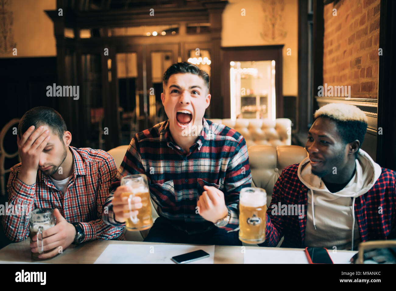 Watching TV in bar. Two happy young men drinking beer and gesturing while sitting in bar Stock Photo