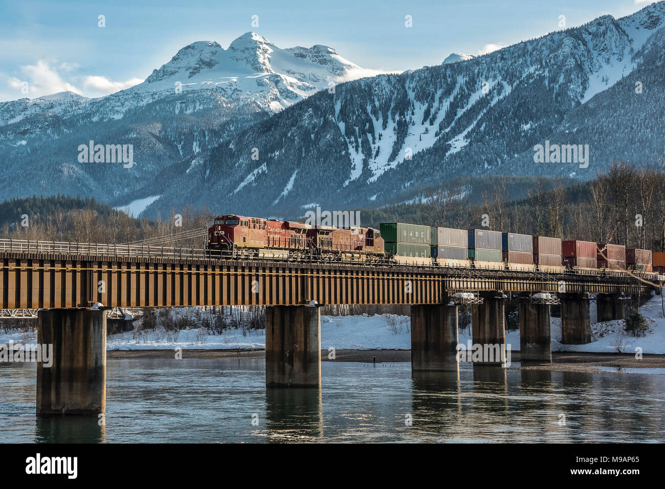 CPR loco 9351 leads intermodal train in winter over Columbia River bridge before Mt Begbie at Revelstoke BC Stock Photo
