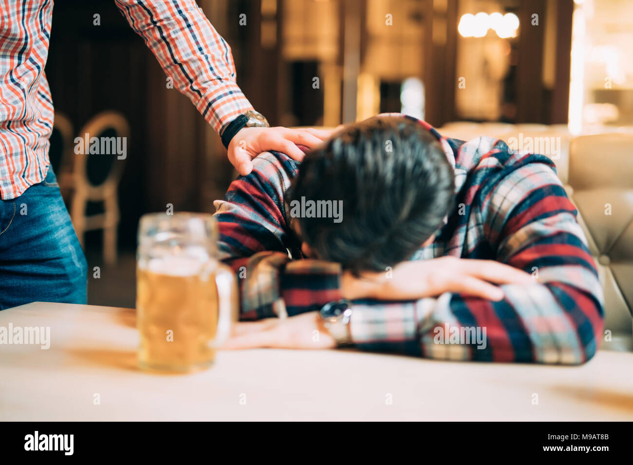people, leisure, friendship and party concept - man with beer waking his drunk friend sleeping on table at bar or pub Stock Photo