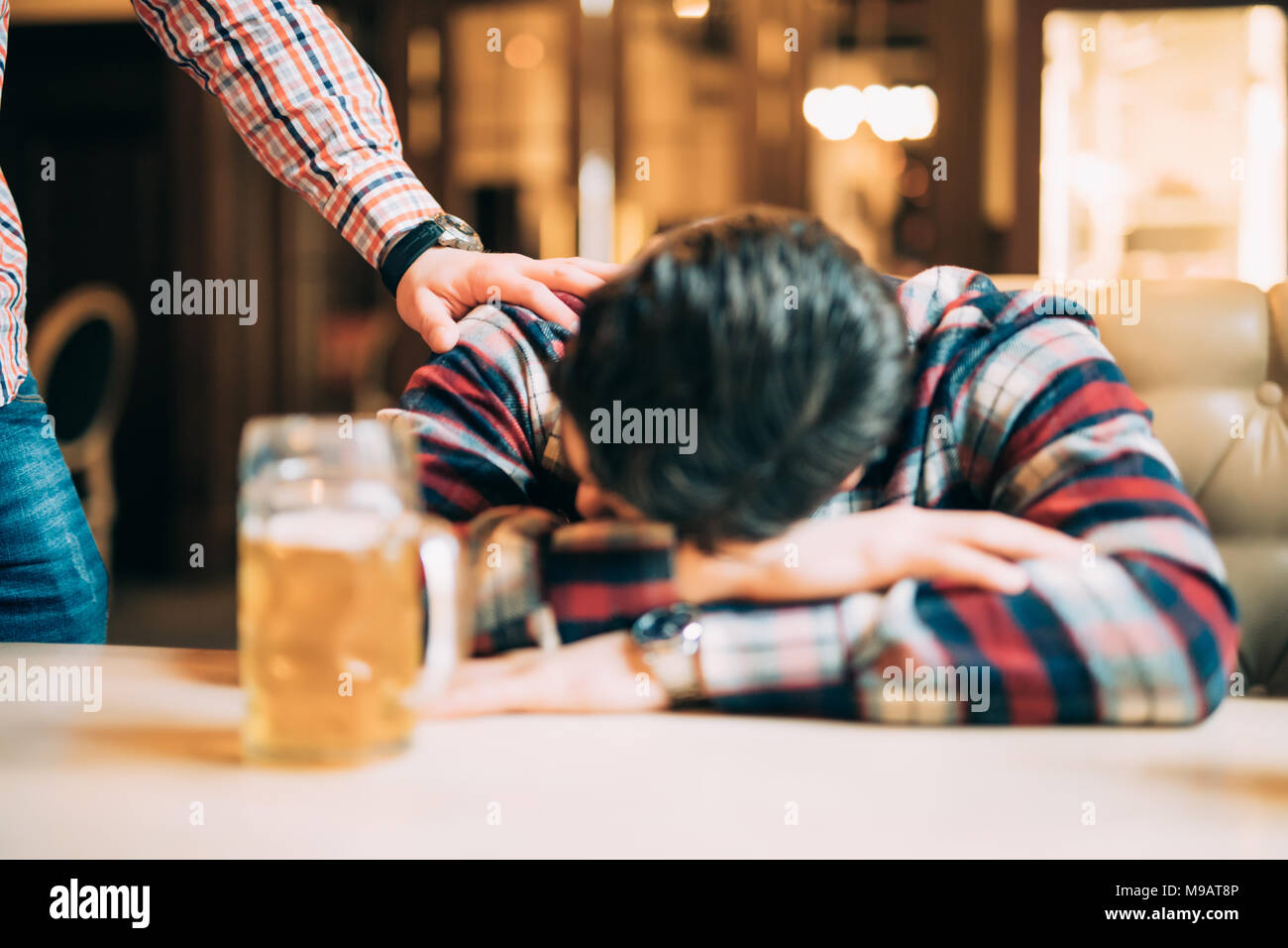 Young man in casual clothes is sleeping near the mug of beer on a table in pub, another man is waking him up. Stock Photo