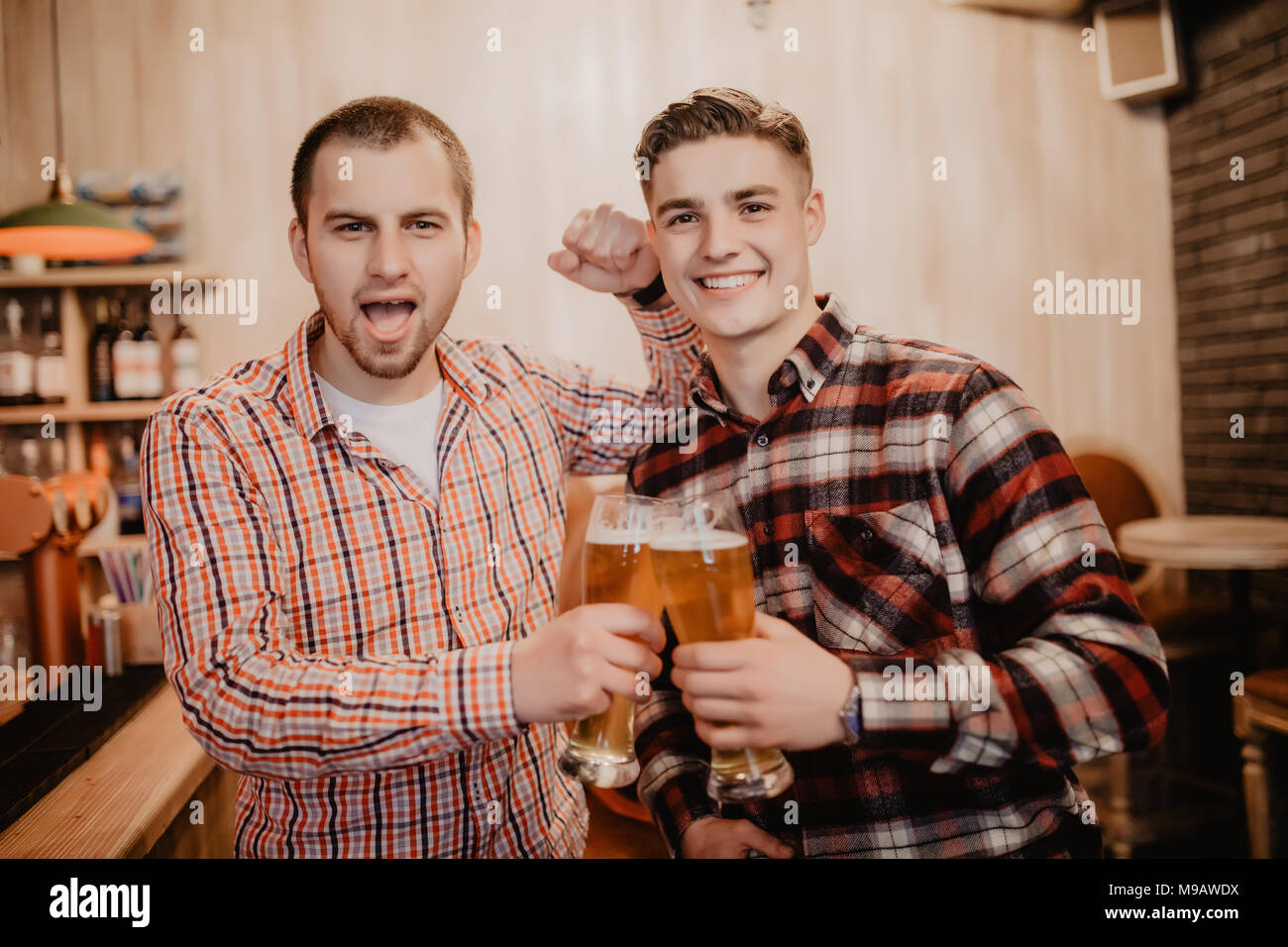 Two handsome young men in casual wear holding glasses with beer and looking away while standing at the bar counter Stock Photo