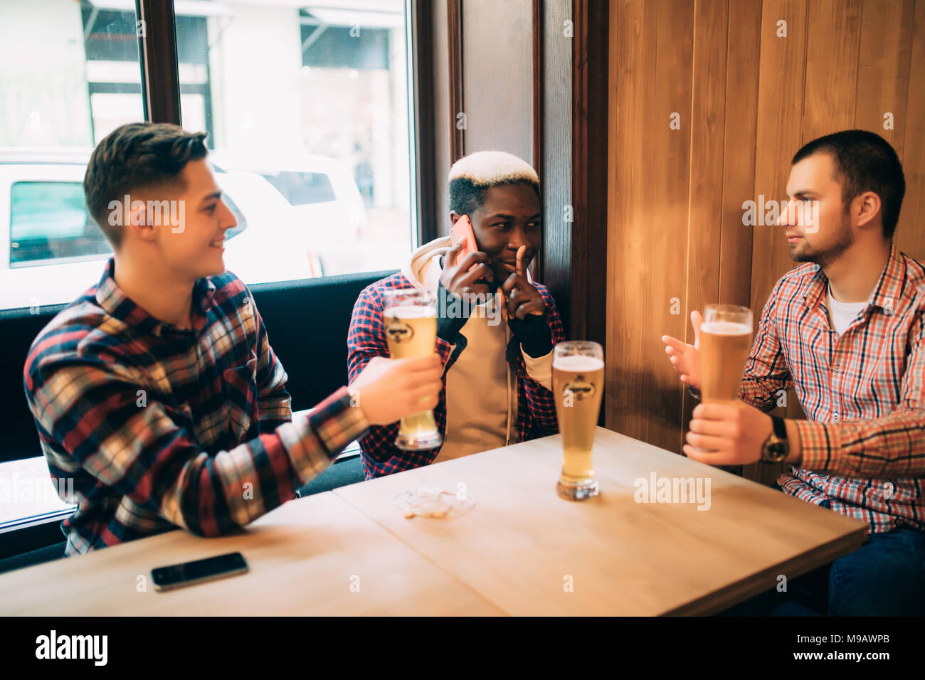 Two male friends in bar are drinking beer and communicating while one is talking on phone and asking for silence. Stock Photo