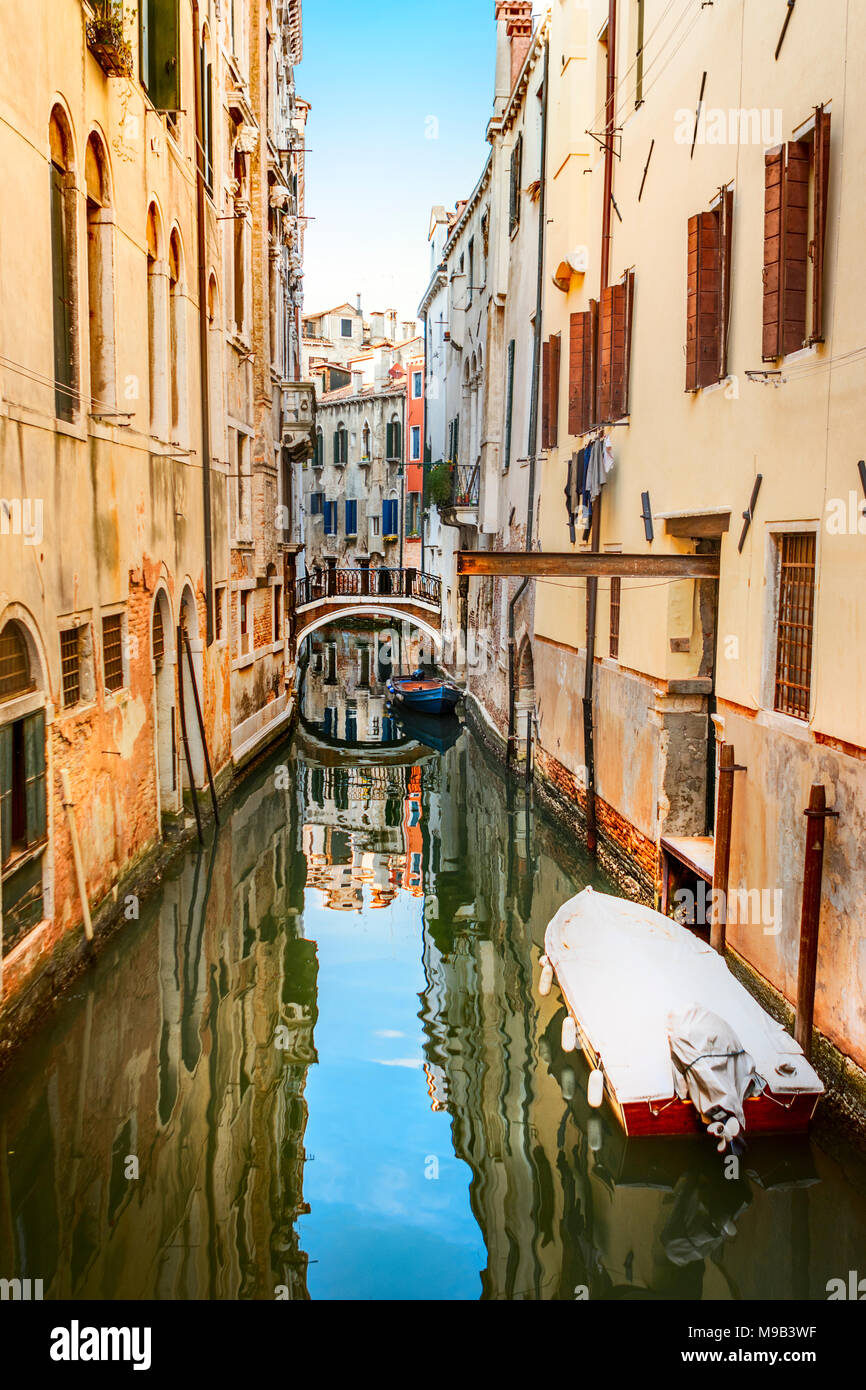 Narrow backstreet canal with reflection of sky in water, a small bridge and colourful buildings with shuttered windows, and boats Stock Photo