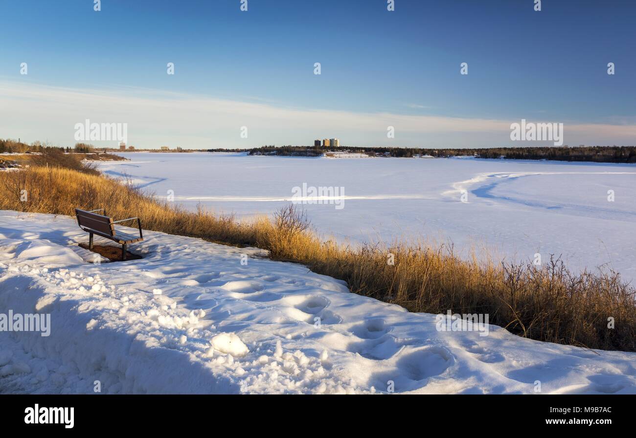 Isolated Park Bench and Distant Snowy Glenmore Reservoir Scenic Landscape in South Calgary Alberta on early Springtime Sunny Afternoon Stock Photo
