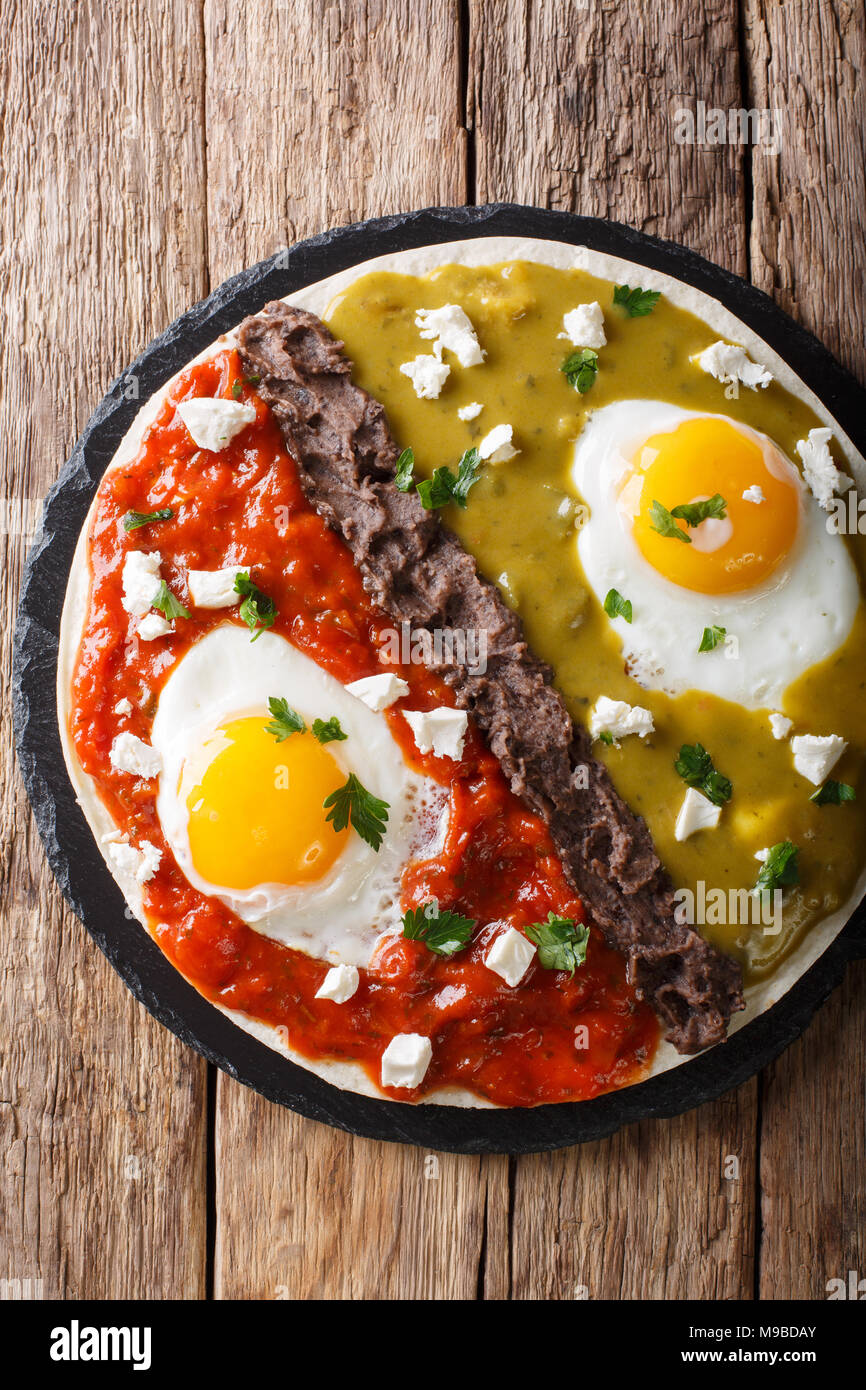Mexican fried huevos divorciados eggs with salsa verde and roja, cheese, black beans on a tortilla close-up on a table. Vertical top view from above Stock Photo