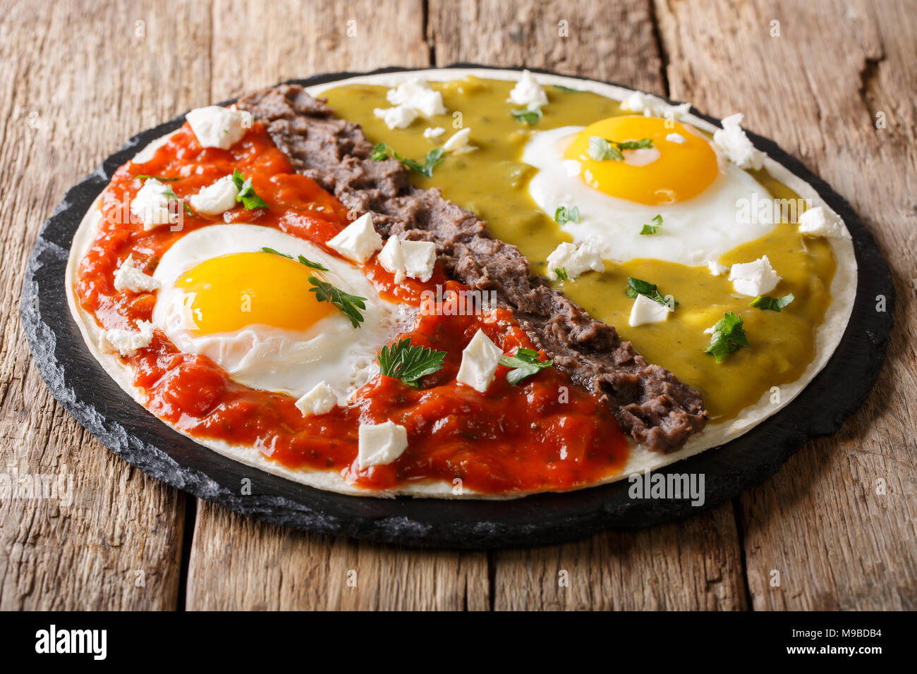 Mexican fried huevos divorciados eggs with salsa verde and roja, cheese, black beans on a tortilla close-up on a table. horizontal Stock Photo