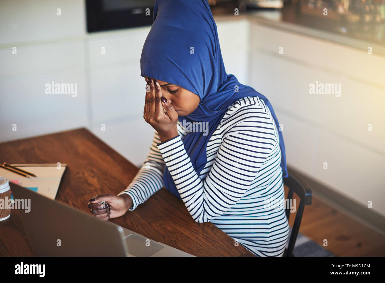 Stressed out young Arabic female entrepreneur wearing a hijab sitting at her kitchen table using a laptop while working from home Stock Photo