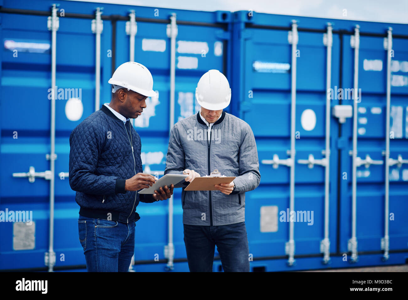 Two engineers comparing inventory lists using a tablet and a clipboard while standing by freight containers in a shipping yard Stock Photo