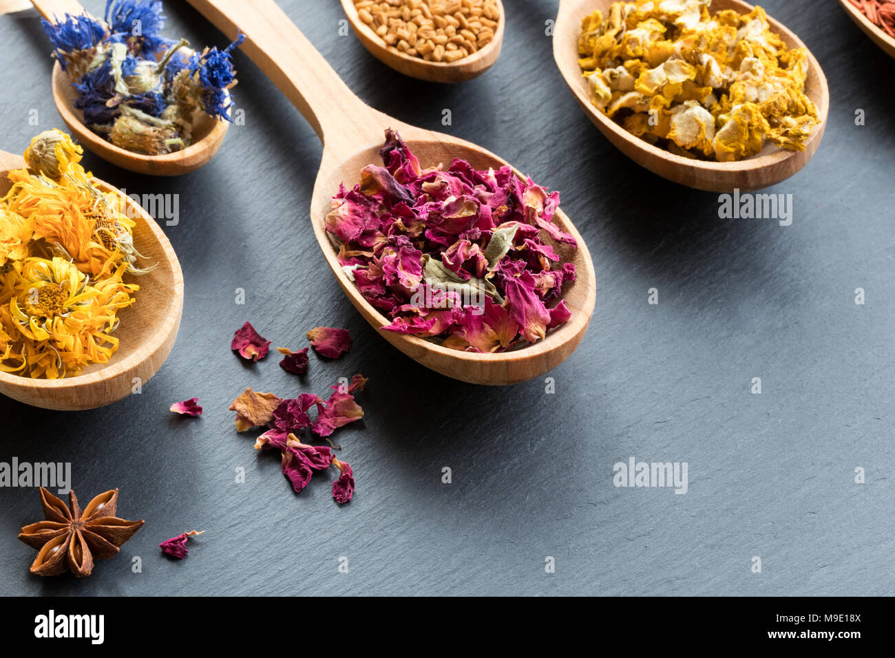 Herbs on wooden spoons on a dark background with copy space. Dried calendula, cornflower, rose petals, fenugreek seeds, mullein, star anise. Stock Photo