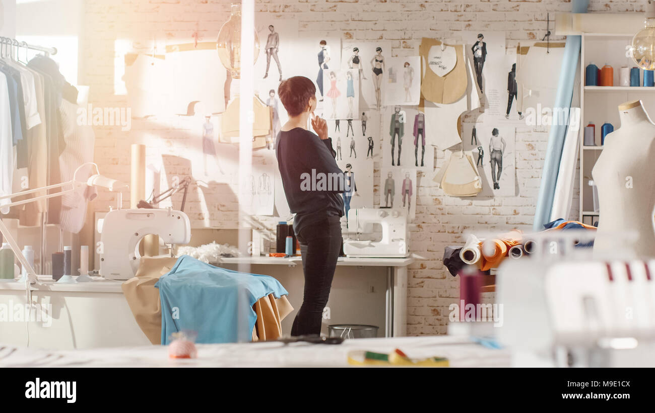 Female fashion, designer,  Looking at Drawings and Sketches that are Pinned to the Wall Behind Her Desk. Studio is Sunny. Personal Computer Stock Photo