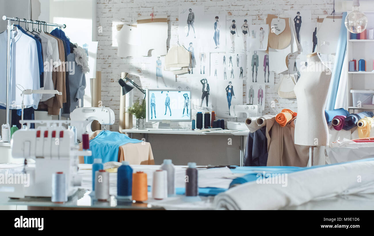 Shot of a Sunny Fashion Design Studio. We See Working Personal Computer, Hanging Clothes, Sewing Machine and Various Sewing Related Items on the Table Stock Photo