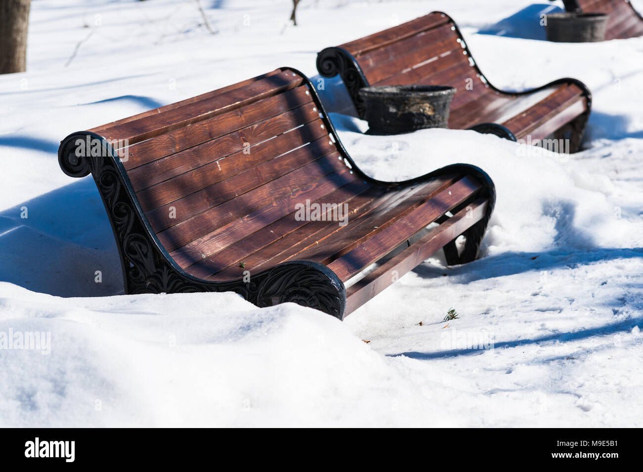 Deep white snow around brown metal and wood benches in a public park on a sunny day of early spring Stock Photo
