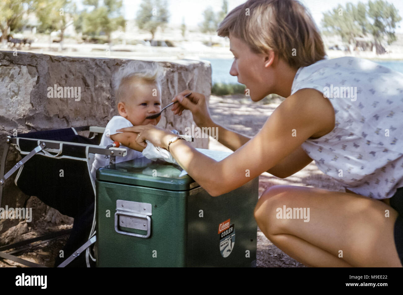 Attractive young woman feeding a 7 to 8 month old baby girl at a campsite in USA in the 1950s with a large green metal camping cooler Stock Photo