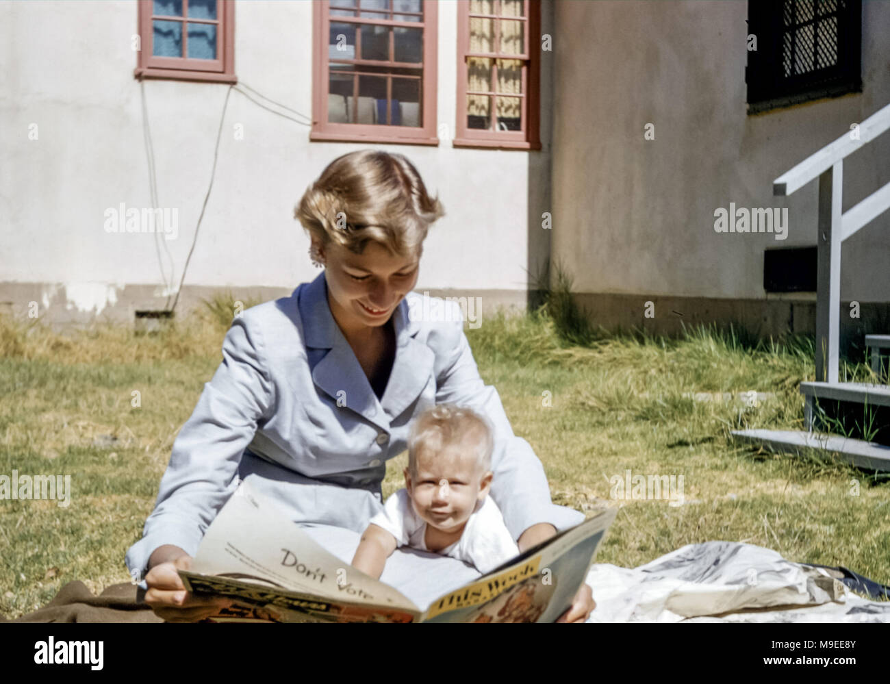 Smiling young woman sitting in a garden with 4 to 5 month old blonde haired baby in sunshine looking at a magazine called This Week. House in California, USA in the 1950s Stock Photo