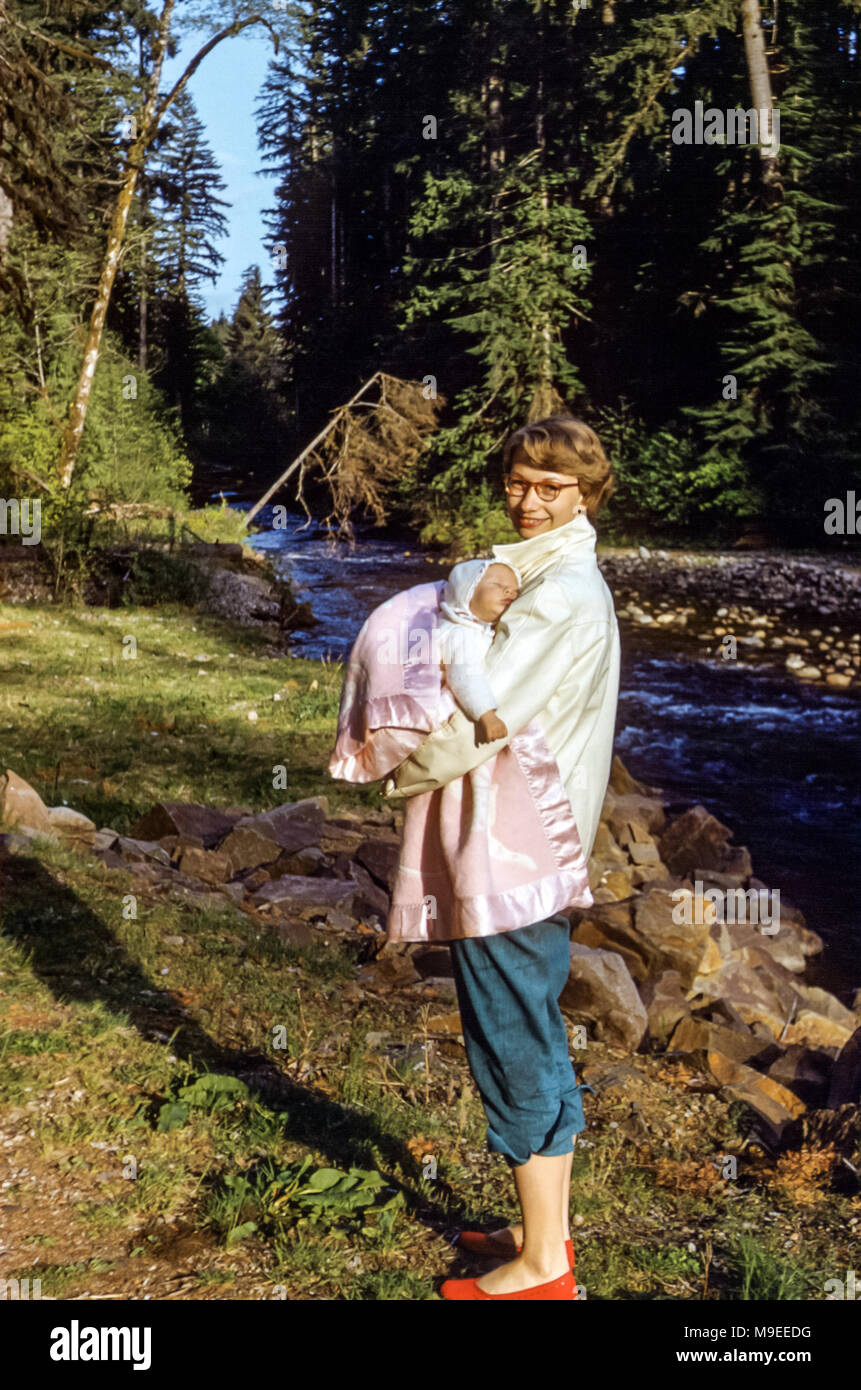 Young smiling woman wearing glasses, shorts and red shoes holding a sleeping baby wrapped in a shawl next to a river, Spirit Lake, Washington state, USA in 1950s Stock Photo