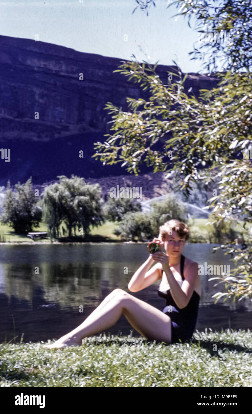 Young woman wearing one piece bathing costume swimsuit sitting on the bank of a lake looking at herself in a pocket mirror, in USA in 1950s Stock Photo