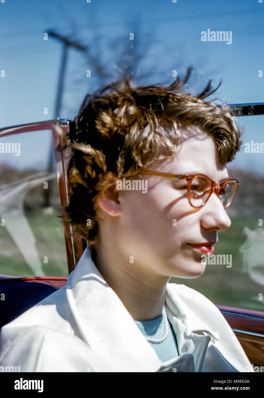 Close up of young woman wearing glasses and red lipstick with hair blowing in the wind while driving a red 1952 Plymouth convertible car with the top down travelling on a road in USA in 1950s Stock Photo