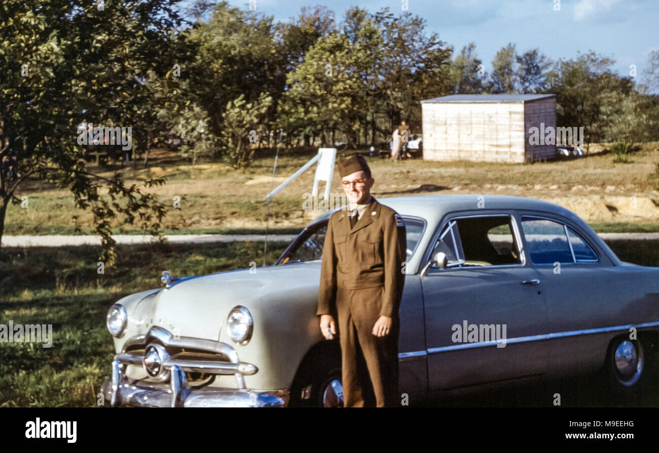 Young man wearing glasses in army military uniform during US draft service standing next to a 1949 Ford sedan car at Fort Dix, New Jersey, USA in 1950s Stock Photo
