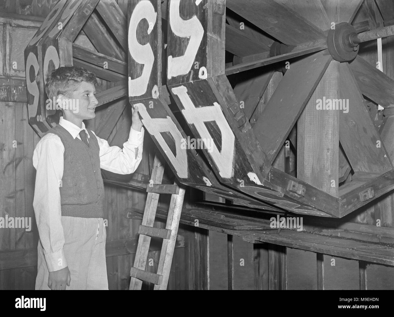 Boy inside an old wooden cricket scoreboard hut, dwarfed by the massive score 'wheels', c. 1950. The wheels were turned by hand by the scorers inside to display the correct number in a window at the front of the hut to the players and spectators at the sports venue. These days most sport scoreboards are electronic and digital displays - a vintage nineteen fifties photograph Stock Photo