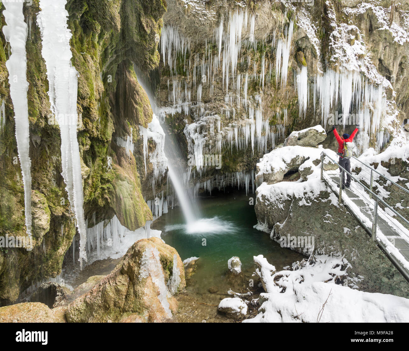 Happy celebrating winning success man standing on bridge with arms raised up above his head in enjoying icicles and waterfall on winter hike with rais Stock Photo