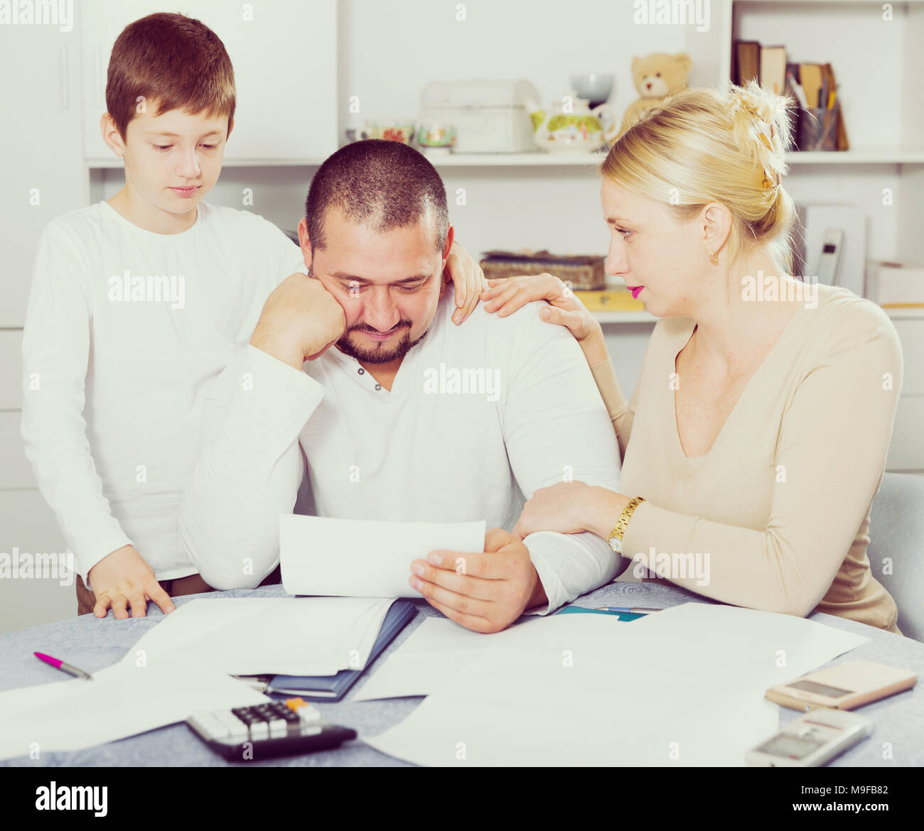 Chagrined man sitting at home table with papers while family soothing ...