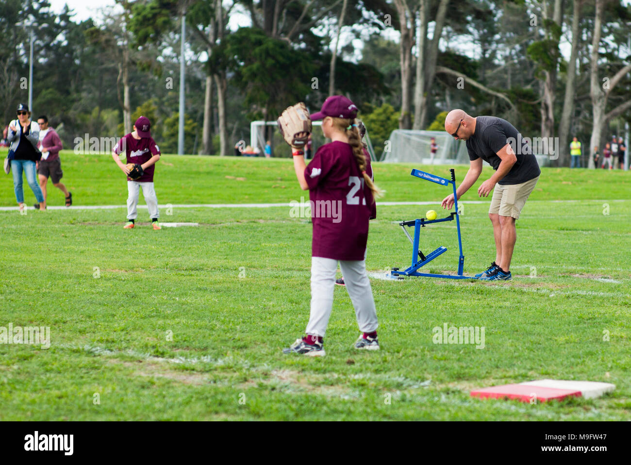 A adult uses a pitching machine following though with pitching arm action at junior softball. Stock Photo