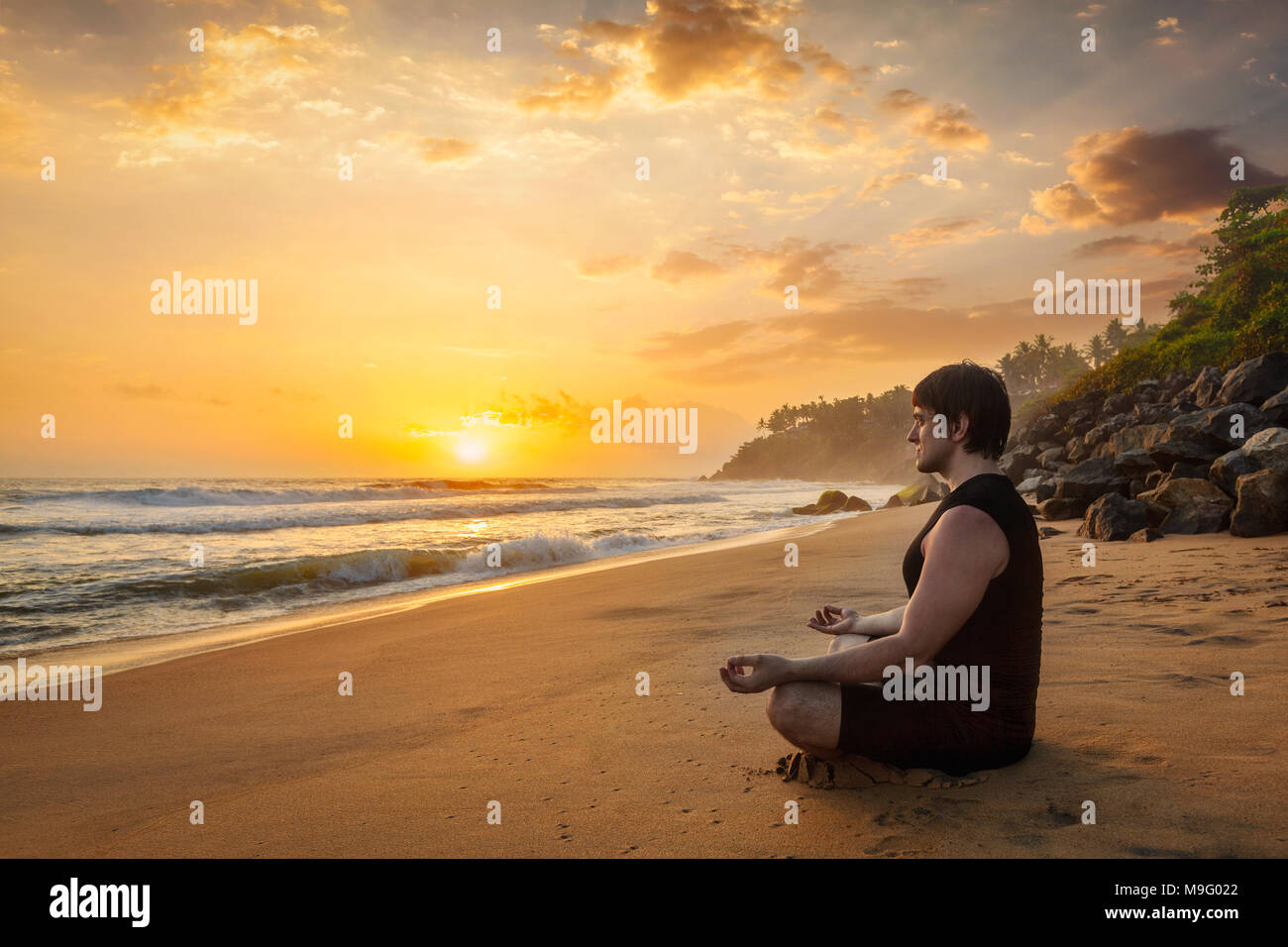 Young sporty fit man doing yoga meditating on tropical beach Stock Photo